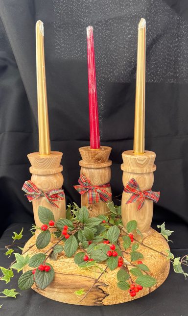 Three candles, two yellow and one red, on a wooden holder decorated with bows and greenery.